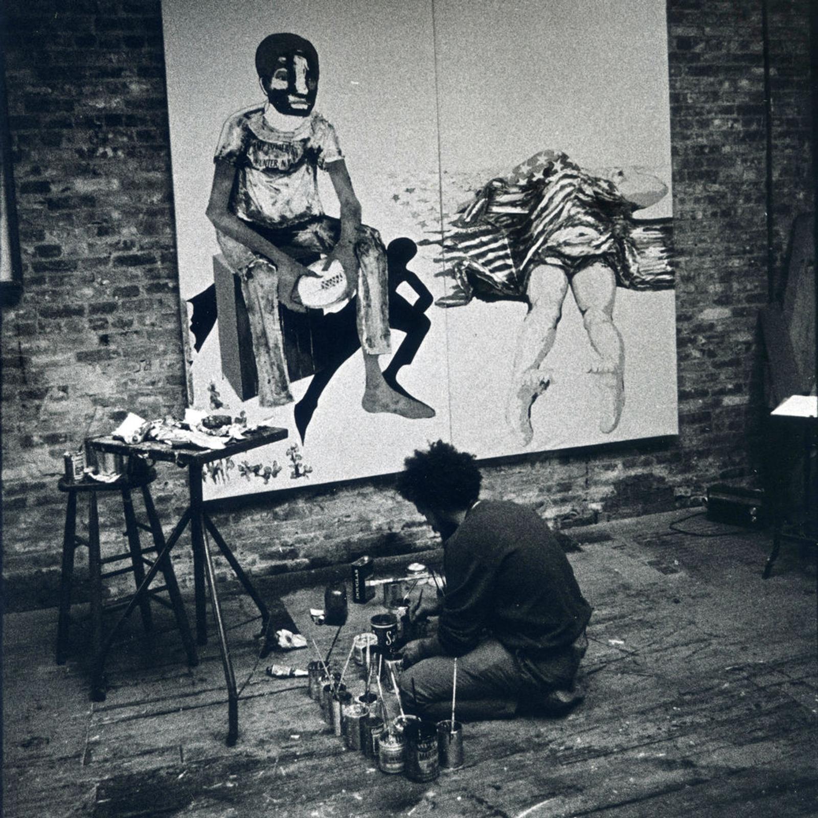 Black-and-white archival photograph of Benny Andrews in his studio, seen from behind as he works facing the painting No More Games.