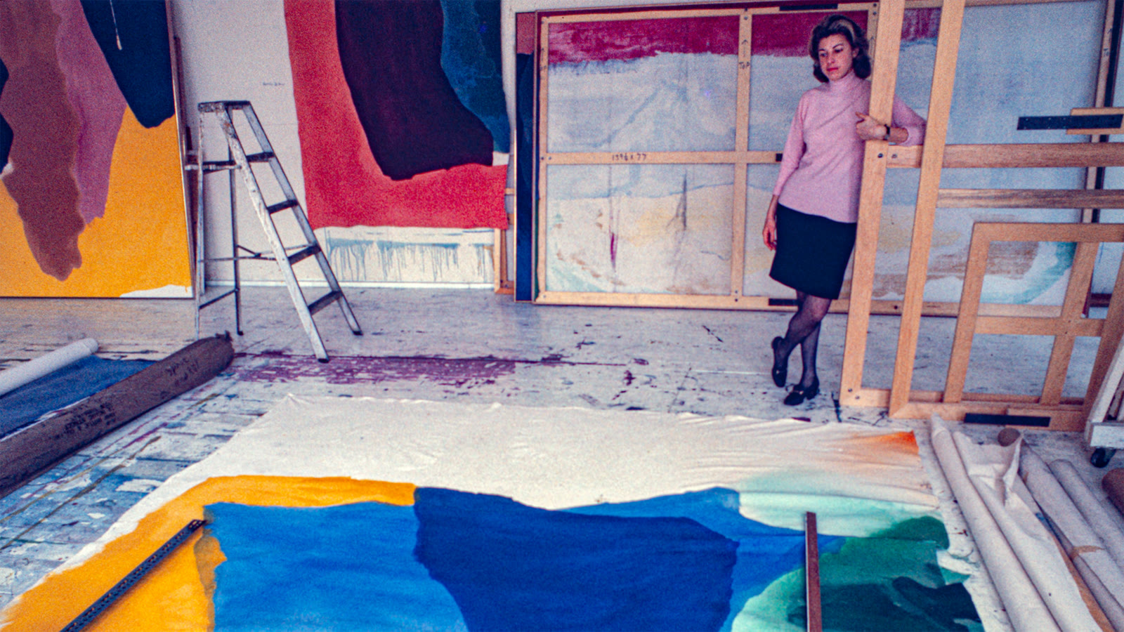 Helen Frankenthaler standing in a colorful, paint-splattered studio surrounded by large abstract canvases.