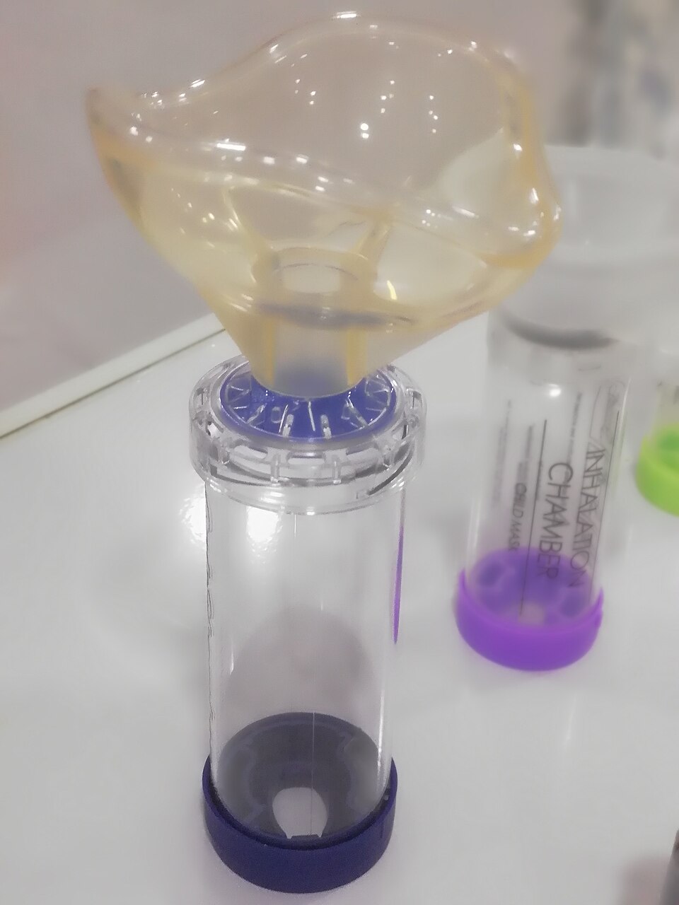 Photograph of a clear metered-dose-inhaler spacer with a blue mouthpiece and attached soft mask.