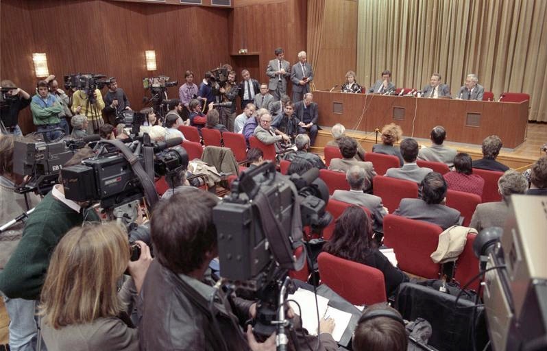 Black-and-white archival photograph of Günter Schabowski speaking behind microphones at a crowded press conference in East Berlin on 9 November 1989.