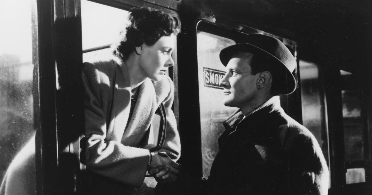 Celia Johnson and Trevor Howard in a black-and-white still from the 1945 film Brief Encounter, standing close beside a train.