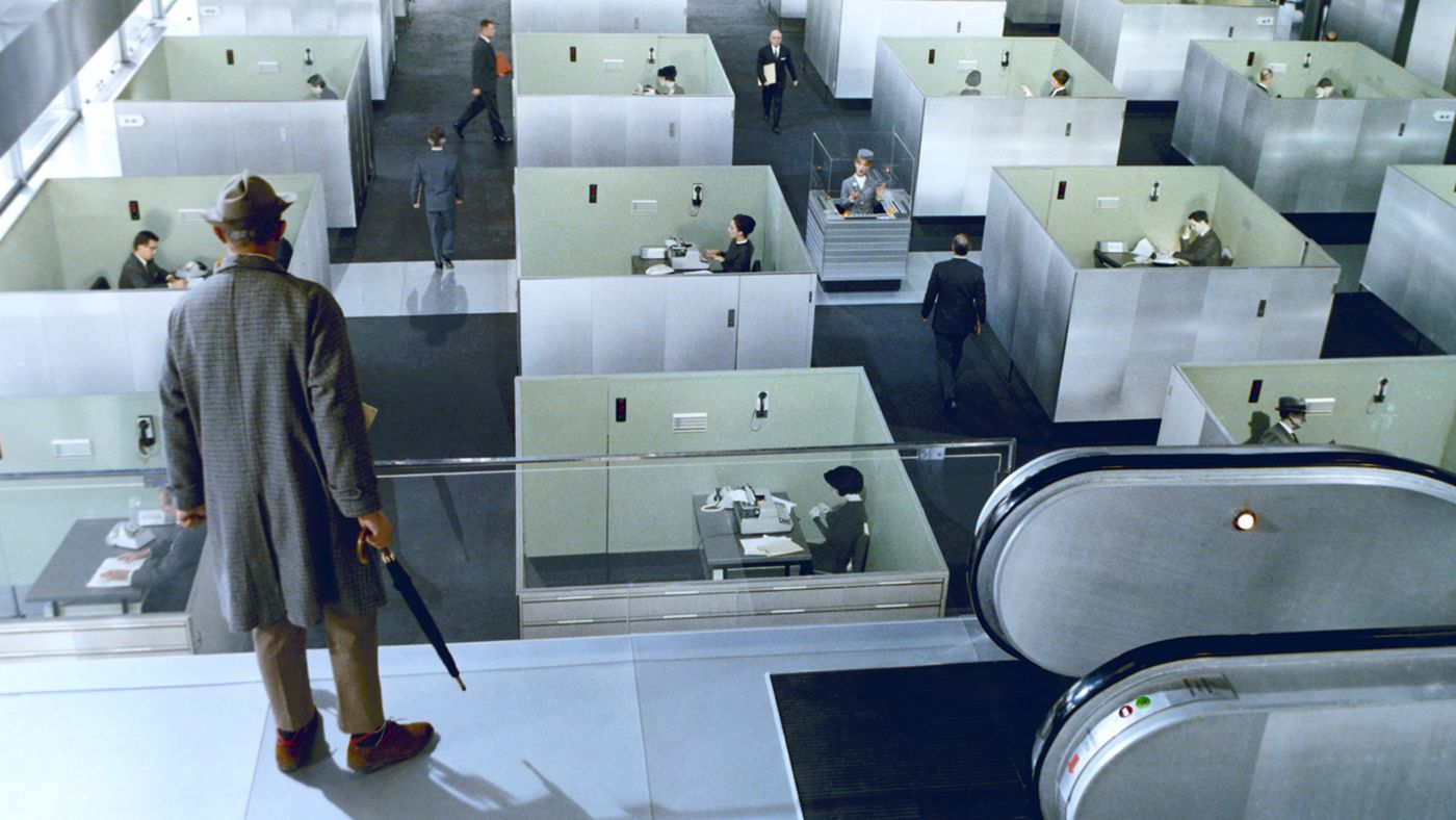A glass-walled modern lobby from Jacques Tati's 1967 film PlayTime, with reflections, chrome, and multiple figures spread across the frame.