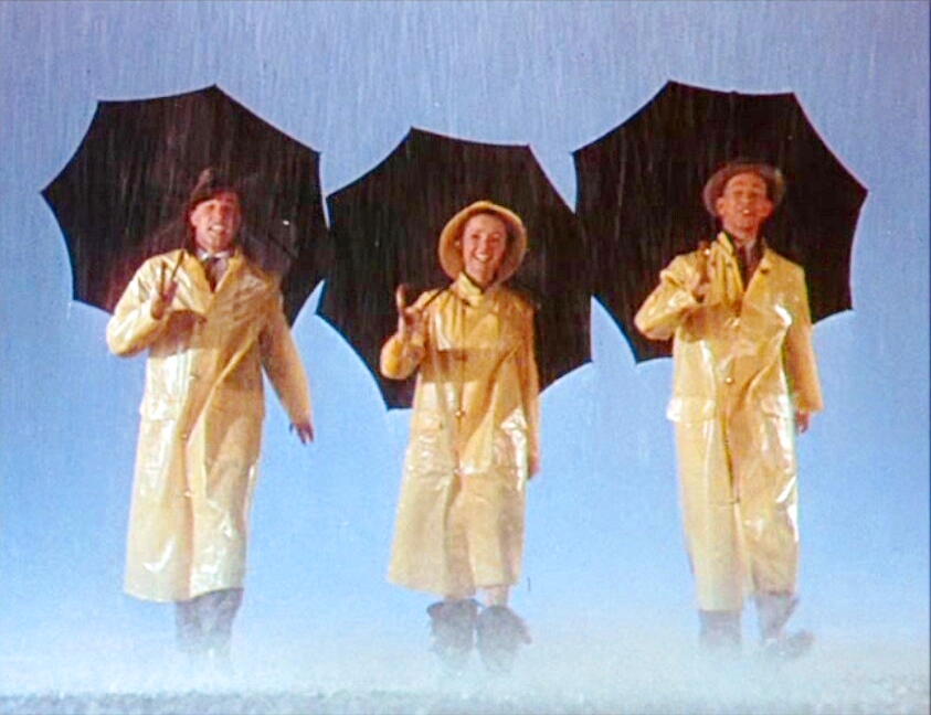 Gene Kelly, Debbie Reynolds, and Donald O'Connor stand in yellow raincoats under black umbrellas in a trailer still from Singin' in the Rain.