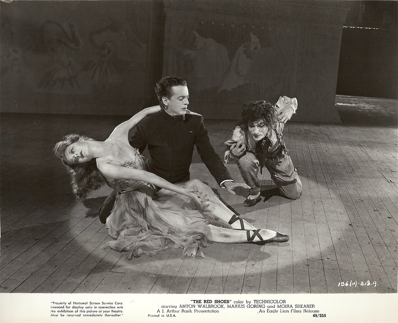 Publicity still from The Red Shoes showing Moira Shearer collapsed on a stage floor while two male performers frame her from either side.
