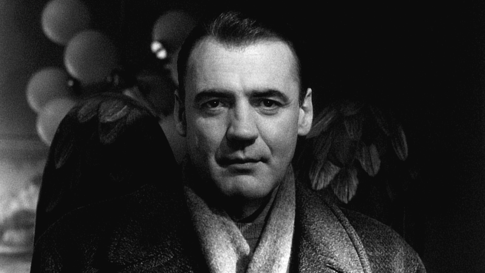 Bruno Ganz as the angel Damiel, facing the camera in a dark coat with feathered wings visible behind him in a black-and-white still from Wings of Desire.
