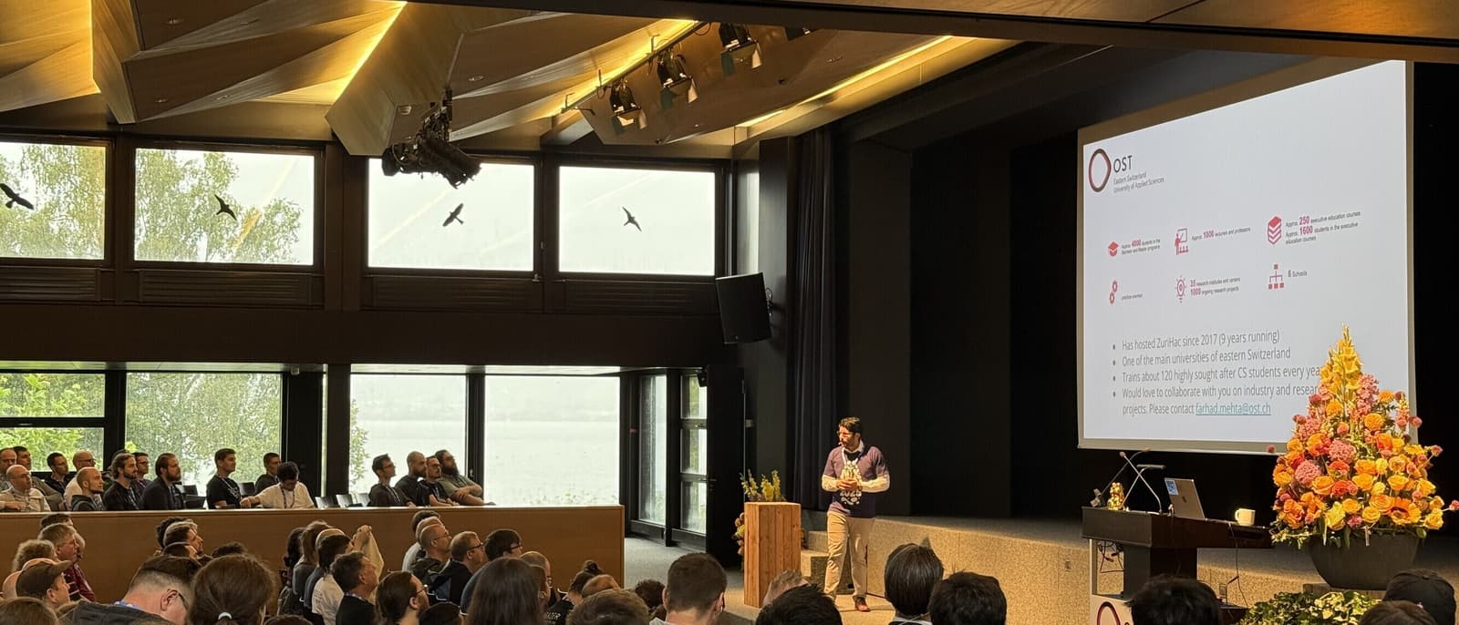 A speaker presenting to a seated audience in a NixCon conference hall at OST, with large windows and a projection screen visible.