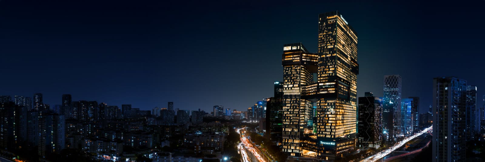 夜色中的腾讯深圳总部大楼群亮着灯，楼下是多车道城市道路。