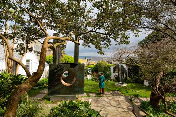 A visitor stands in Barbara Hepworth's sculpture garden in St Ives among dark pierced sculptures, trees, and a view toward the sea.