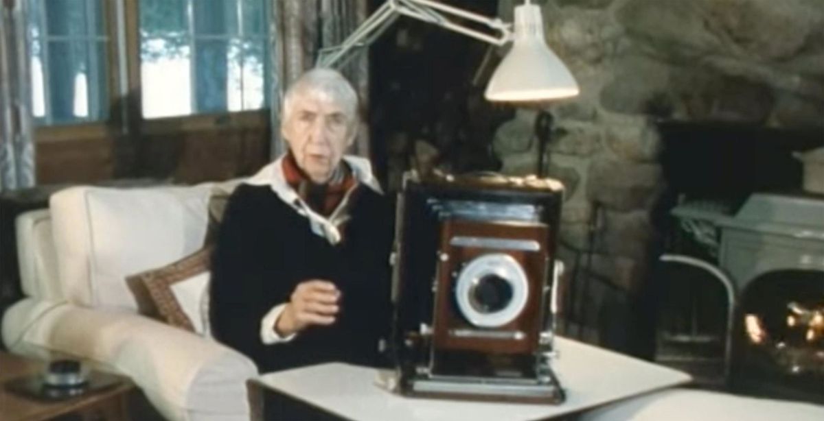 Berenice Abbott seated in an armchair before a fireplace, with a camera resting on a table in front of her.