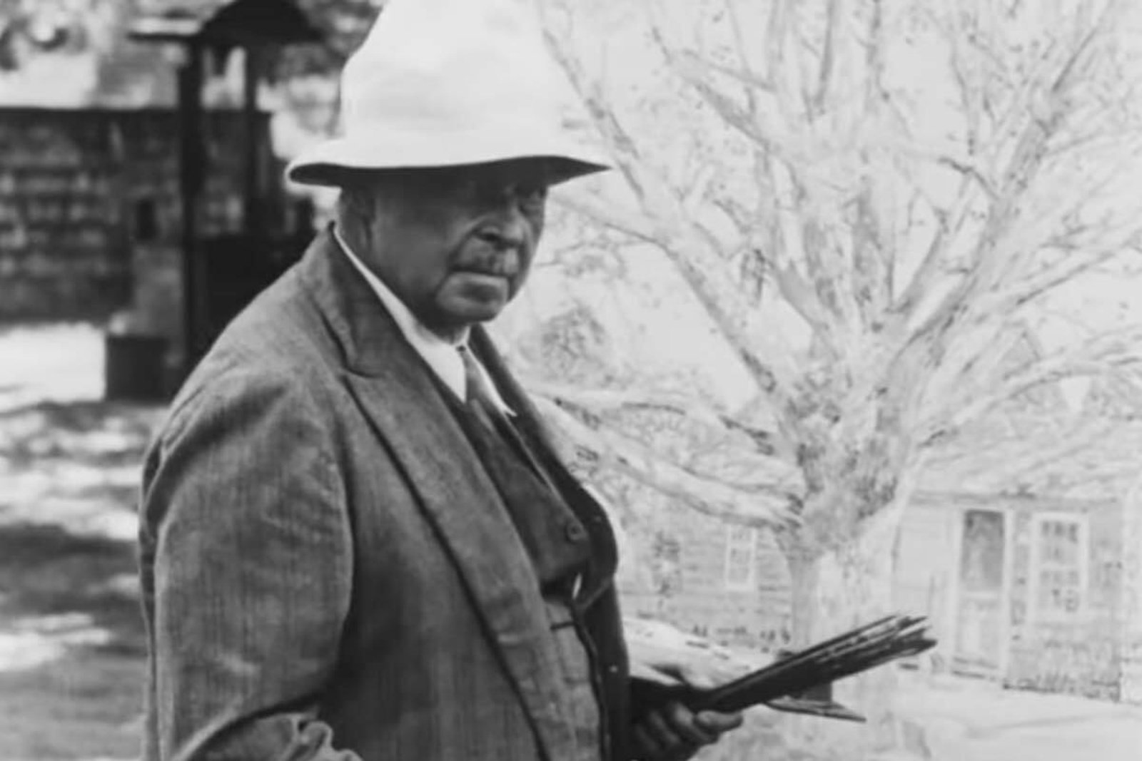 A black-and-white archival photograph of Childe Hassam outdoors in a hat and jacket, holding painting materials.