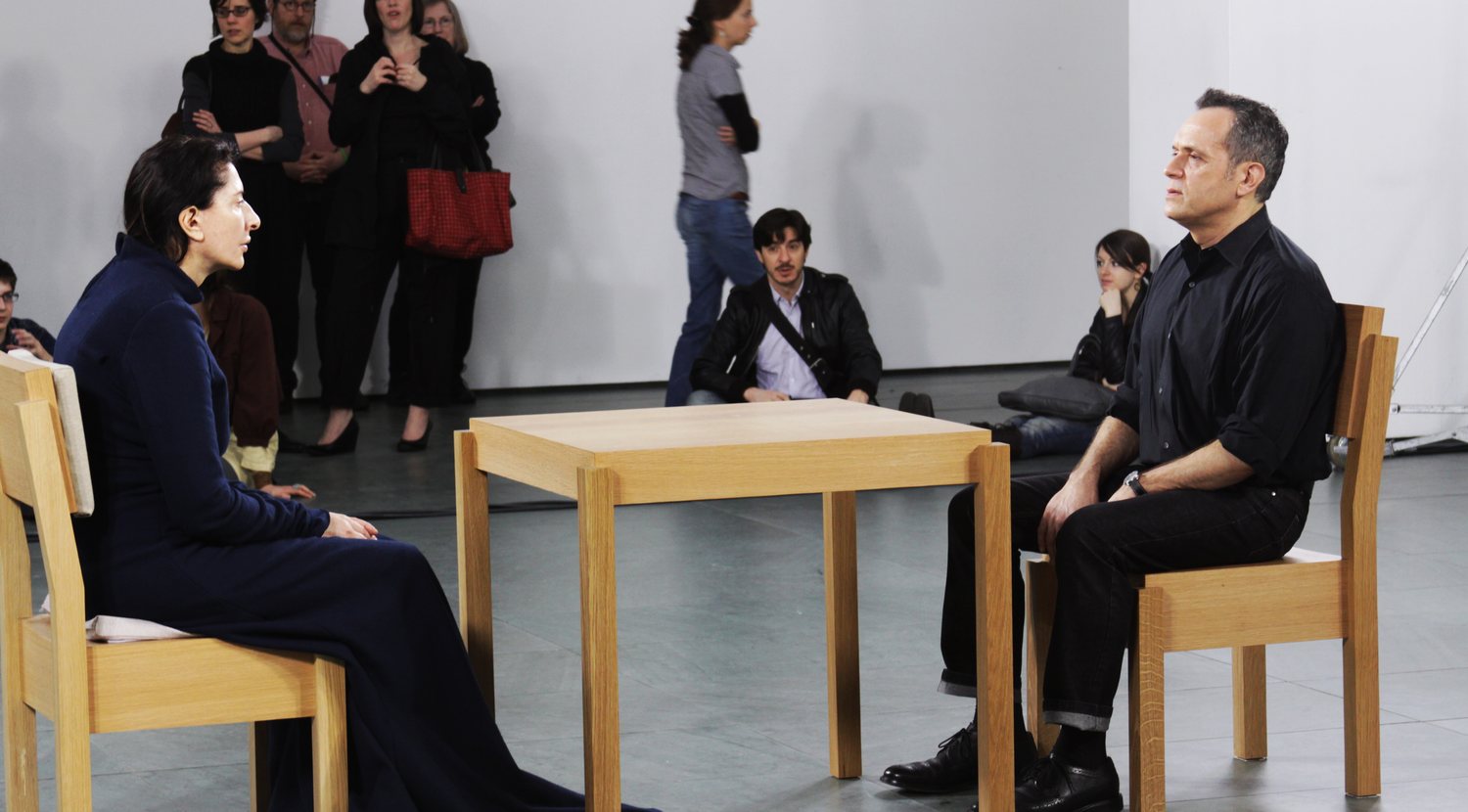Marina Abramovic seated across from a visitor during The Artist Is Present at MoMA in 2010, with a square table between them and museum visitors gathered around the atrium.