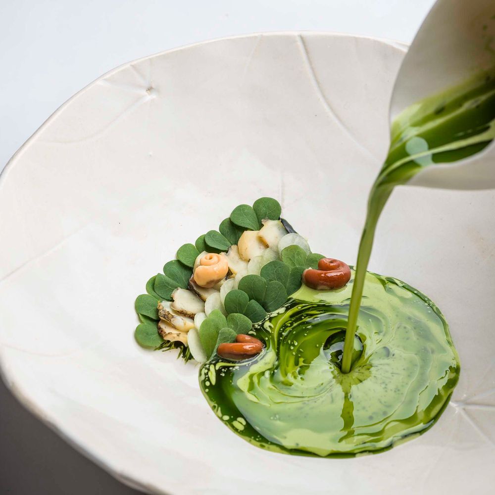 A white bowl at Aponiente holding shellfish and green leaves while a vivid green sauce is poured tableside.