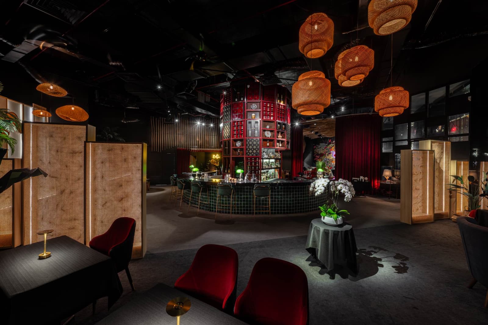 Beta KL's dining room with rattan pendant lights, deep red shelving, dark tables, and softly lit screens around a central bar.