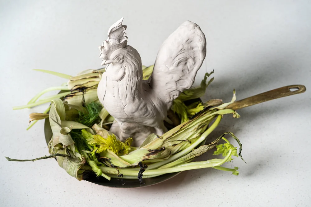 An Esme menu-preview photograph showing a white ceramic rooster standing in a pan with charred vegetables.