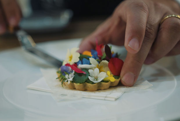A Mirazur tart topped with colorful edible flowers as a hand adjusts it on a white plate.