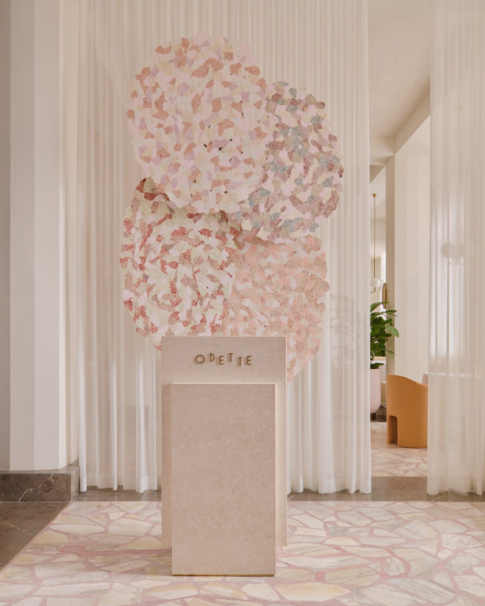 Odette's pale pink reception pedestal beneath a large floral artwork, framed by white curtains and a glimpse into the dining room.