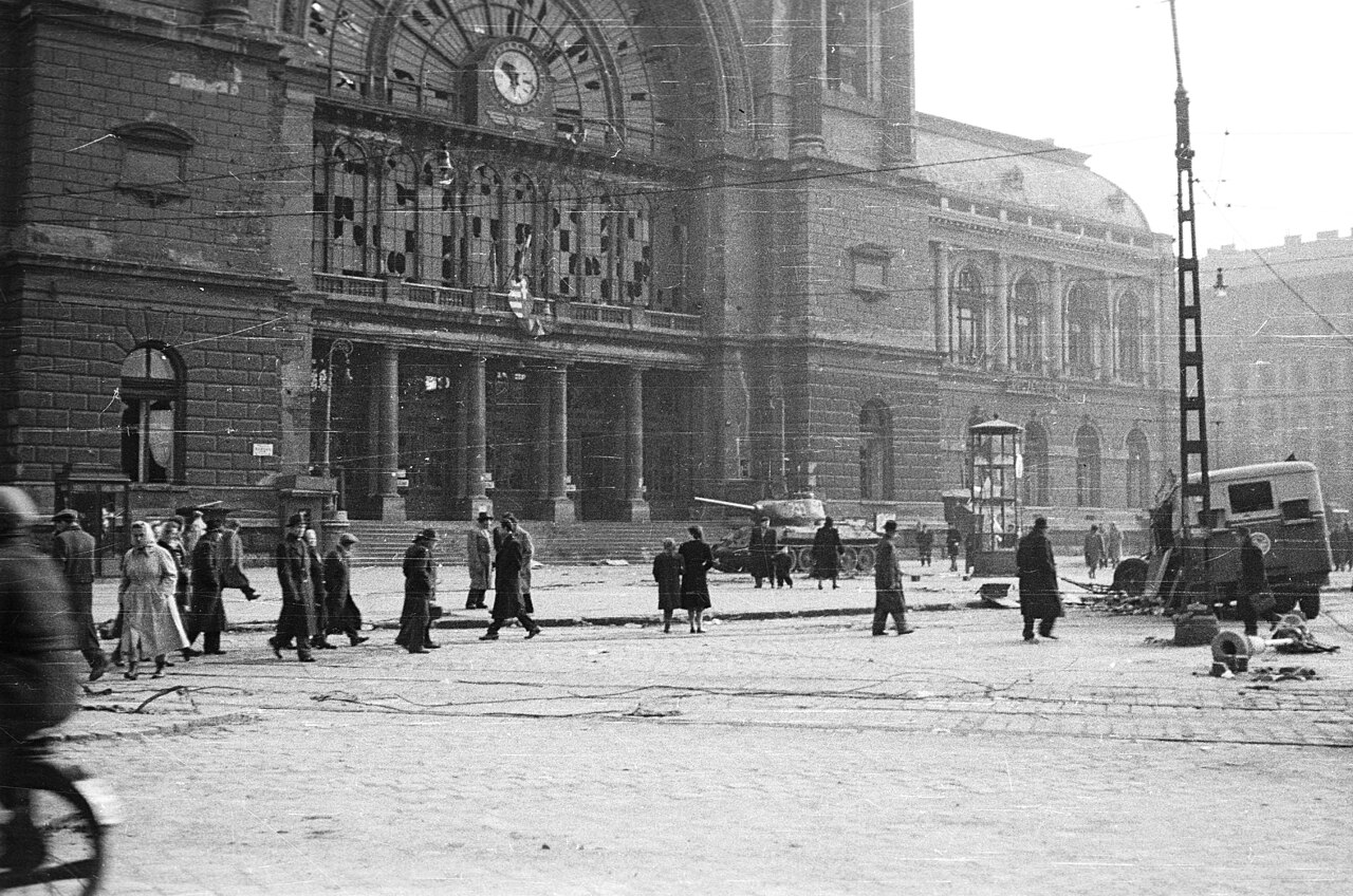 Damaged facade of Keleti railway station in Budapest during the 1956 Hungarian Revolution, with pedestrians crossing the square and a tank visible near the station.