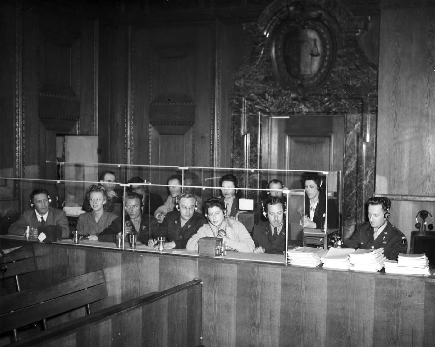 Archival photograph of the interpreters' section at the Nuremberg International Military Tribunal, with translators seated behind glass partitions wearing headsets and working from stacks of papers.