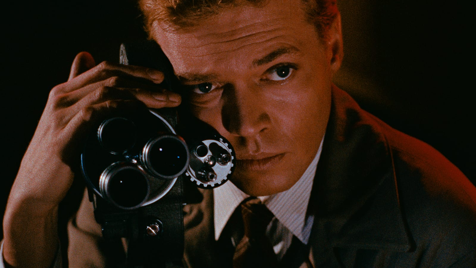 A color still from Peeping Tom shows Mark Lewis holding a film camera close to his face and looking past it with intense concentration.