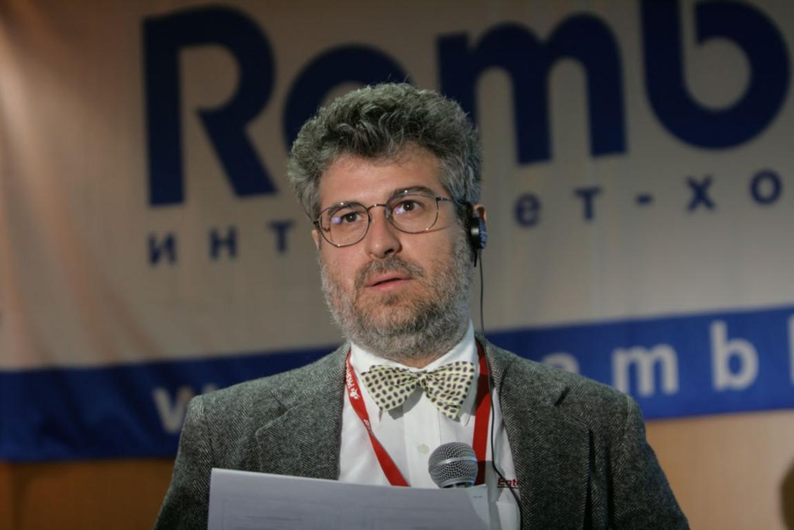 Bruce Momjian speaks at a conference in front of a blurred backdrop, holding papers and a microphone.