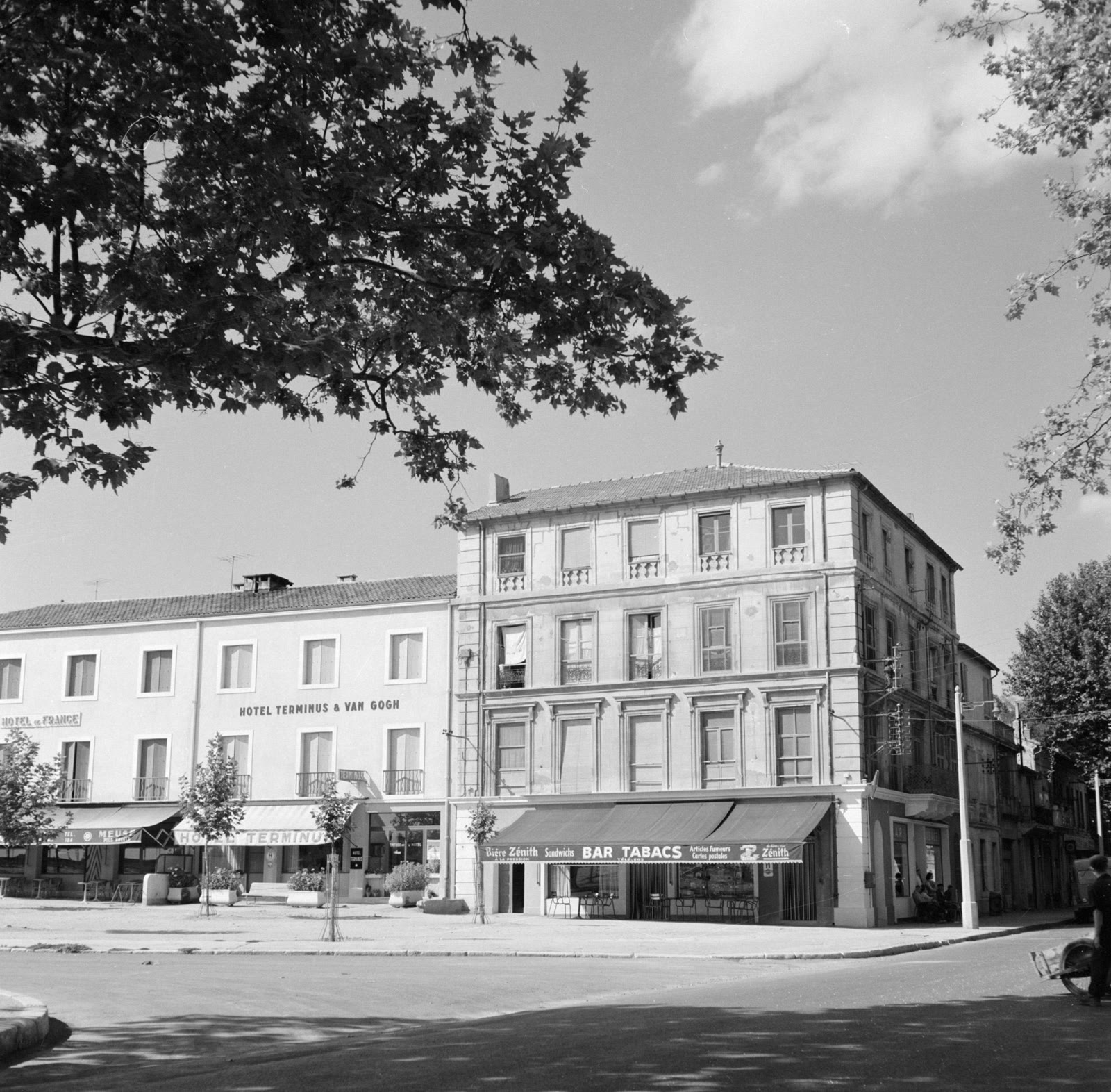An archival black-and-white photograph shows the Place Lamartine corner in Arles associated with Van Gogh's Yellow House, framed by trees and later commercial storefronts.