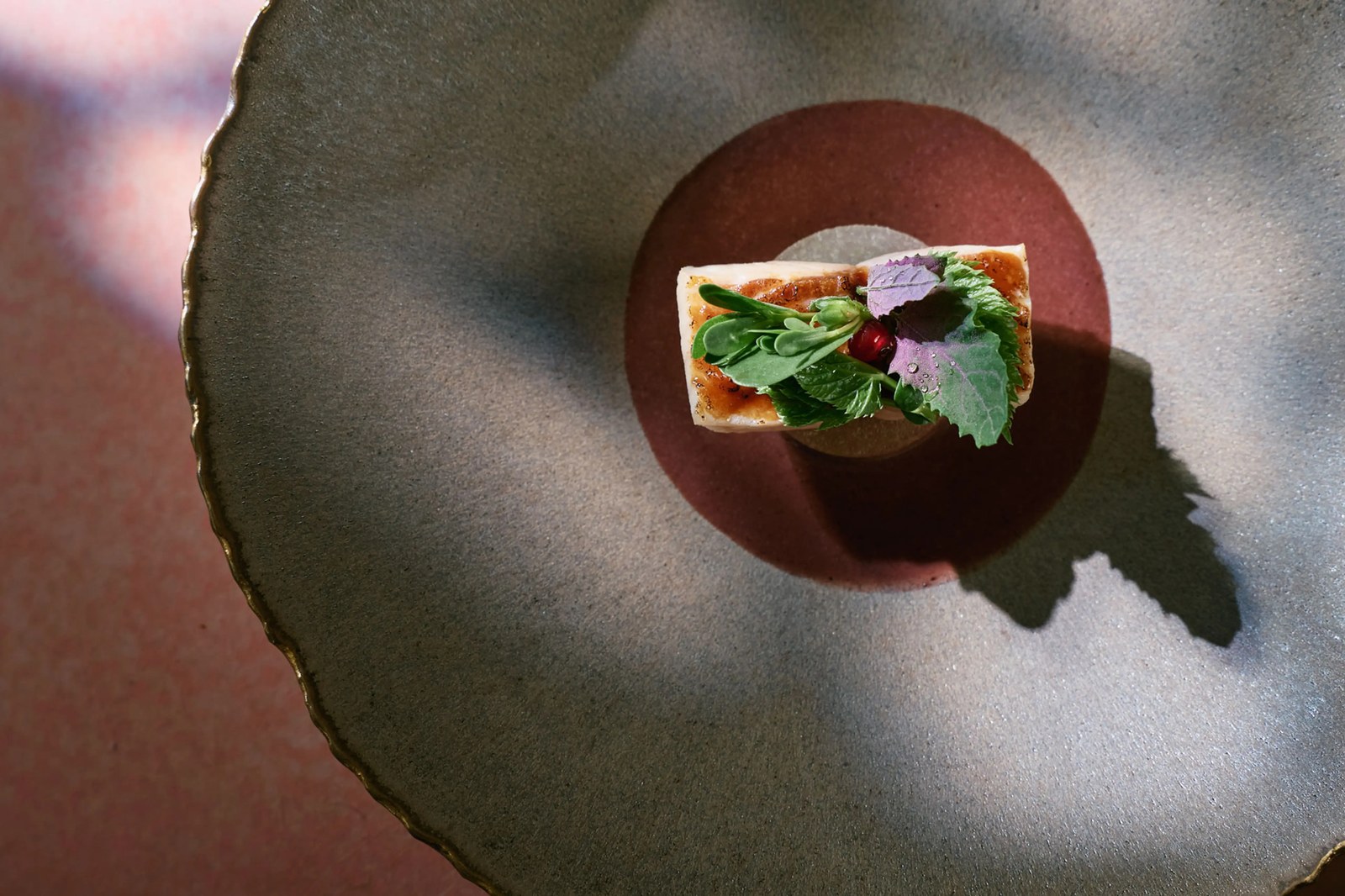 A small rectangular bite topped with wild green and purple leaves sits at the center of a wide dark ceramic plate.