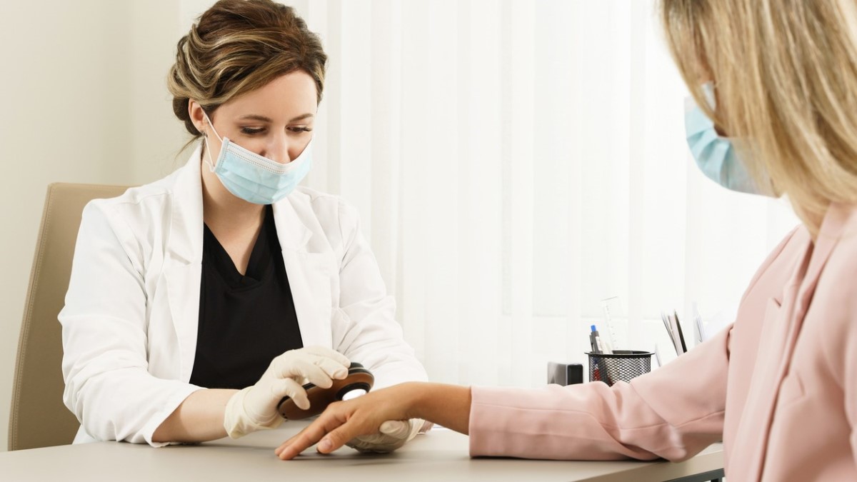 A clinician uses a dermatoscope to examine a patient's hand across a desk in a bright exam room.