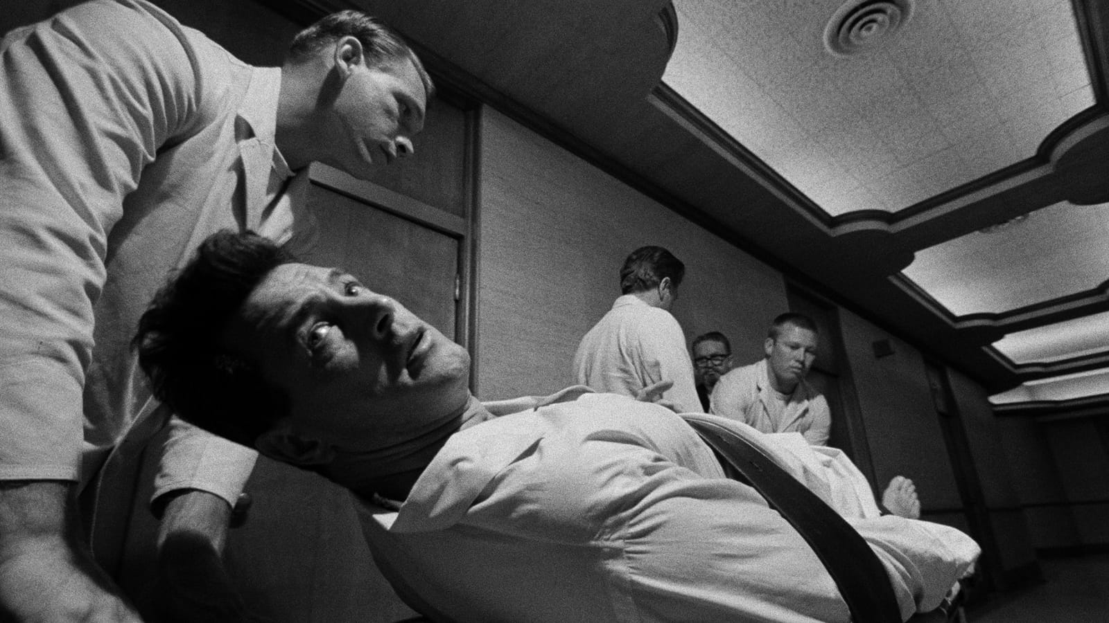 A black-and-white still from Seconds showing a man strapped to a medical table under a canted ceiling while attendants lean over him.