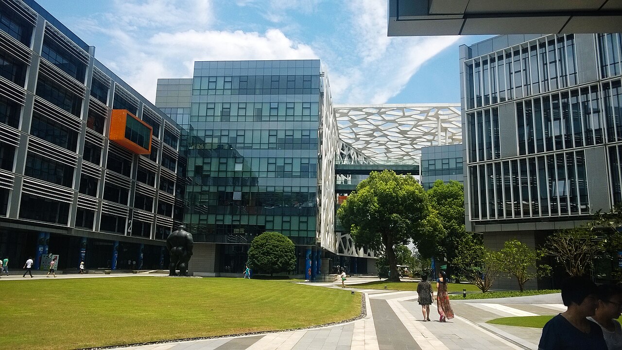 A real photograph of Alibaba Group headquarters in Hangzhou, with glass buildings, trees, and a reflecting pool.