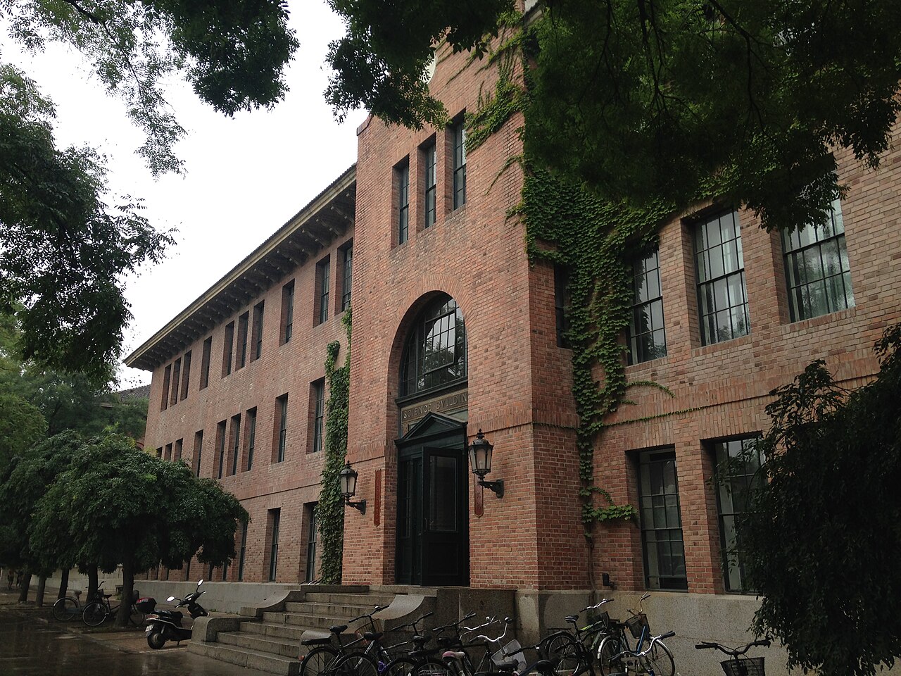 The red-brick Science Building at Tsinghua University in Beijing, photographed from a campus walkway with trees and parked bicycles nearby.