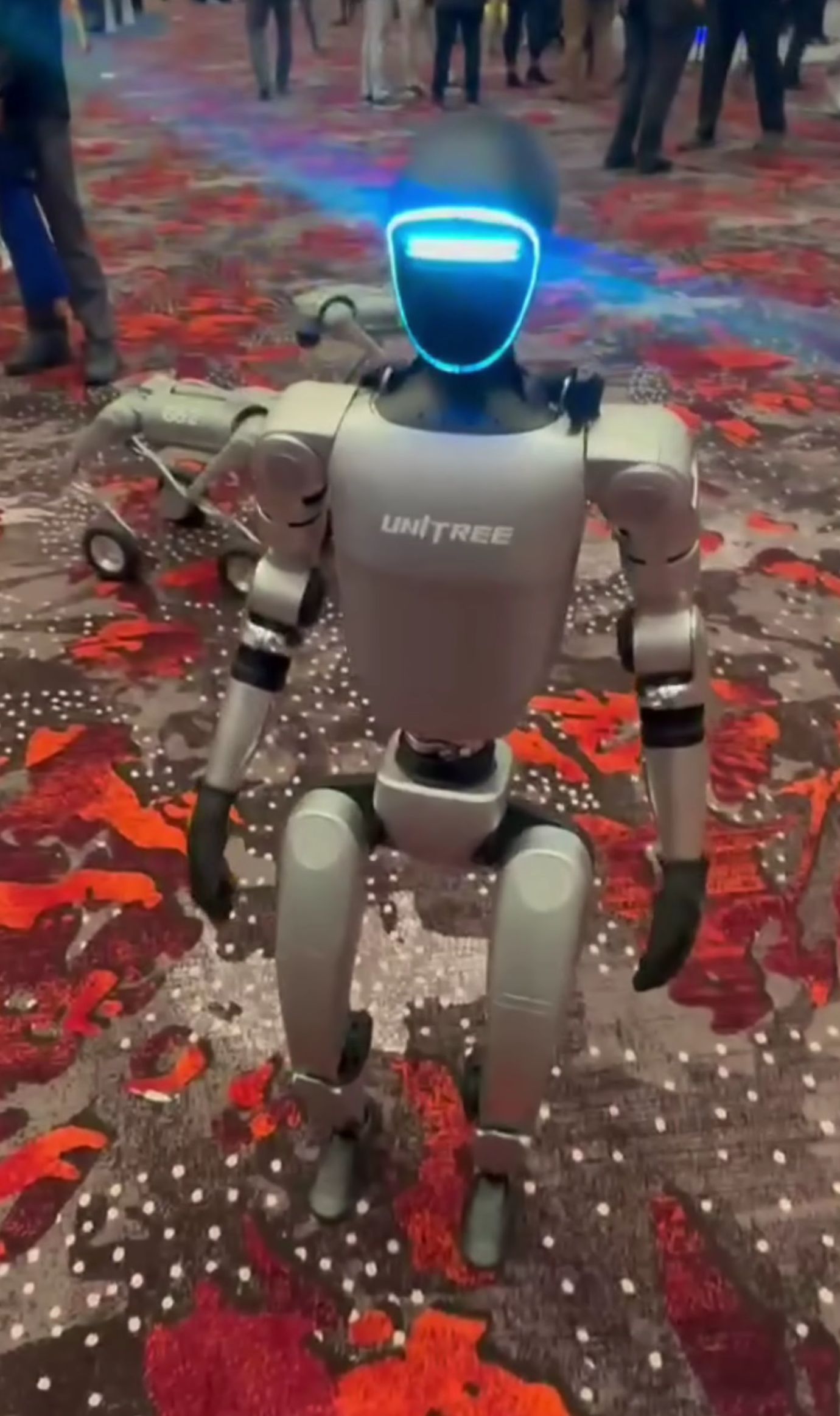 A silver Unitree G1 humanoid robot with a glowing blue face stands on a carpeted expo floor.