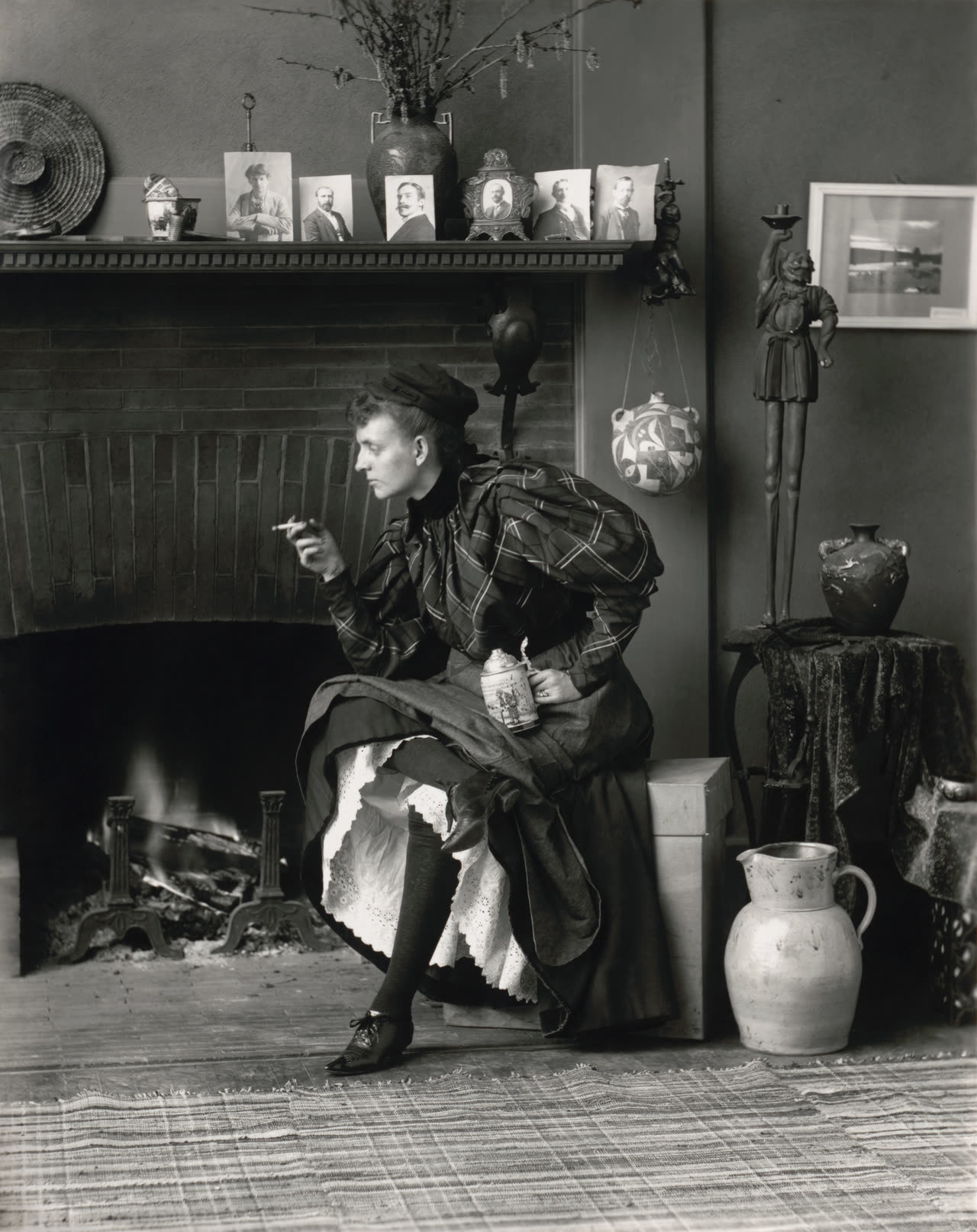 Frances Benjamin Johnston's 1896 black-and-white self-portrait, showing her seated before a fireplace with one leg crossed, holding a cigarette in one hand and a beer stein in the other.