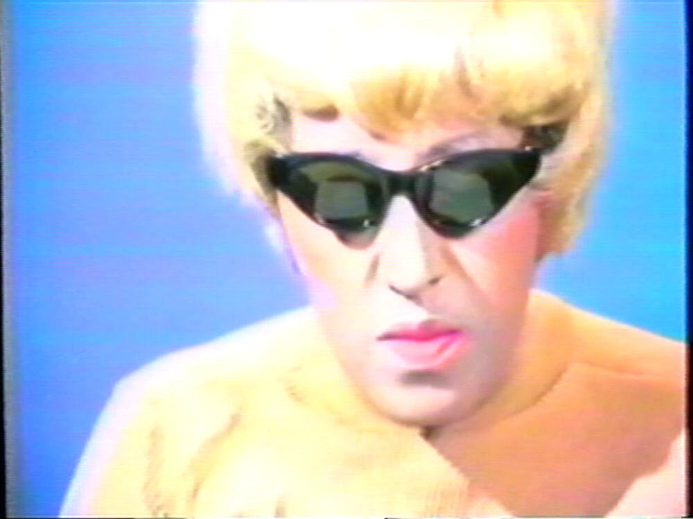 Video still from Howardena Pindell's Free, White, and 21 showing the artist performing a white female character in blond wig and dark glasses against a blue background.