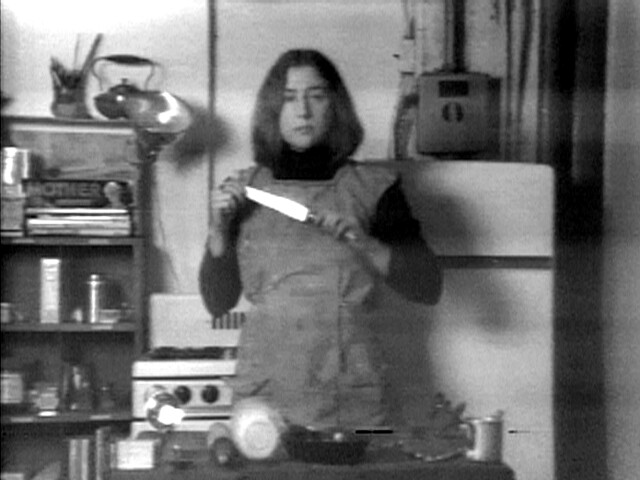 Black-and-white still from Martha Rosler's Semiotics of the Kitchen, showing Rosler in an apron holding a knife in a kitchen.