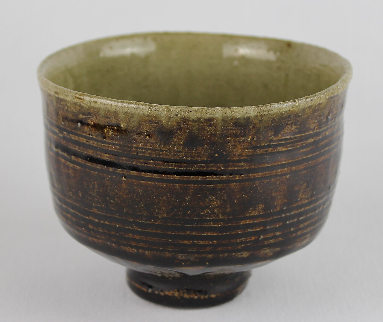 A photographed Shoji Hamada stoneware tea bowl with dark combed exterior bands, pale interior glaze, and a small foot ring.
