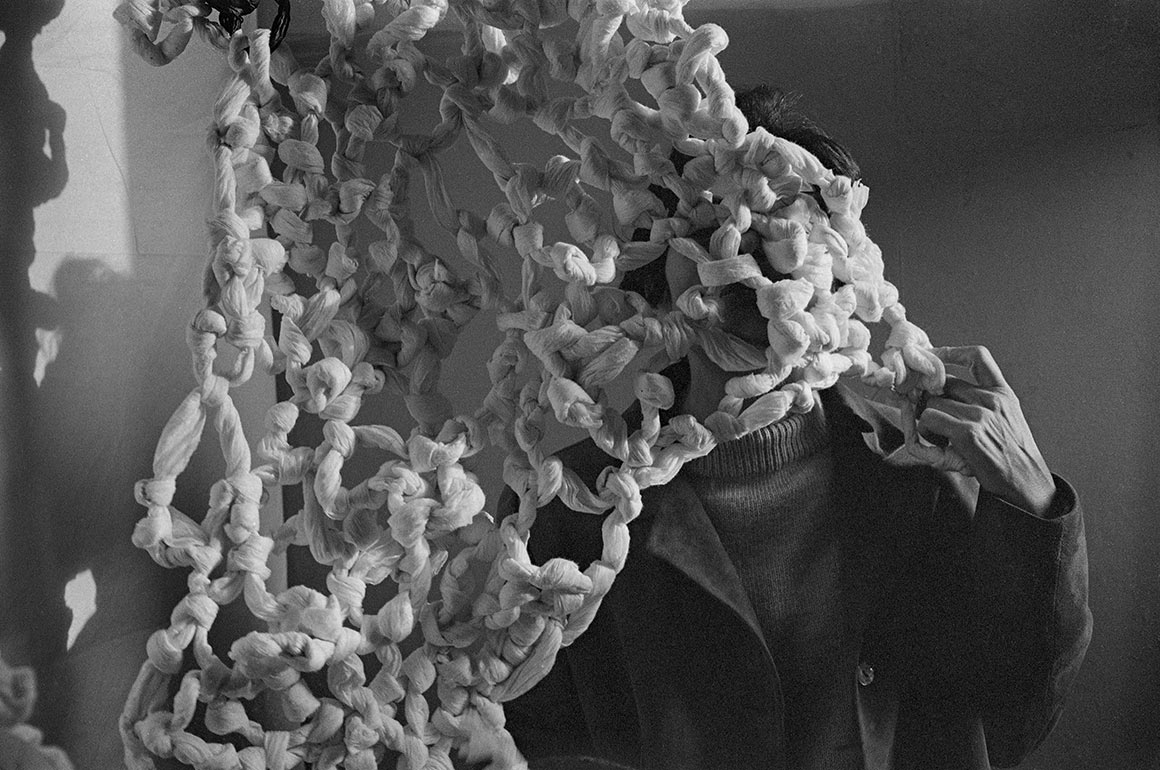 A 1966 black-and-white archival photograph shows Mira Schendel partly hidden behind a hanging tangle of knotted white rice-paper forms.