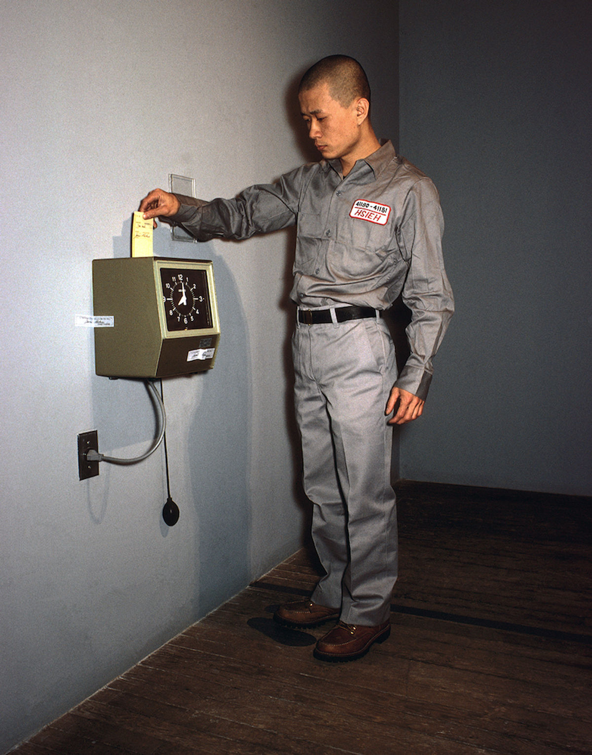 Tehching Hsieh in 1980 standing beside a wall-mounted time clock, reaching toward it in a gray work uniform during One Year Performance 1980-1981.