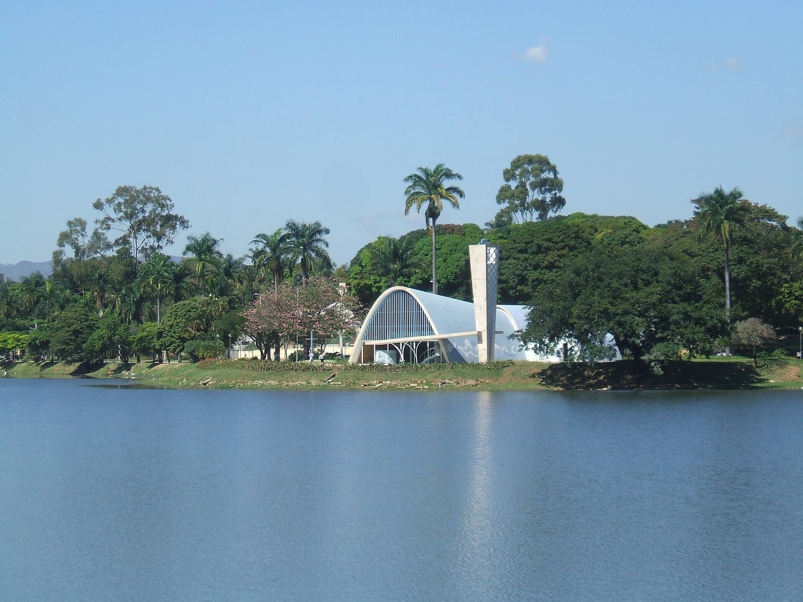 The arched São Francisco de Assis church stands across Lagoa da Pampulha in Belo Horizonte with trees and water in the foreground.