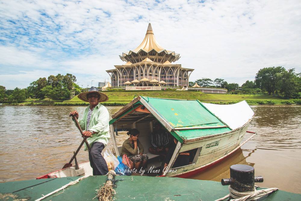 A perahu tambang boatman poles a small covered boat across the Sarawak River with the Sarawak State Legislative Assembly building behind him.