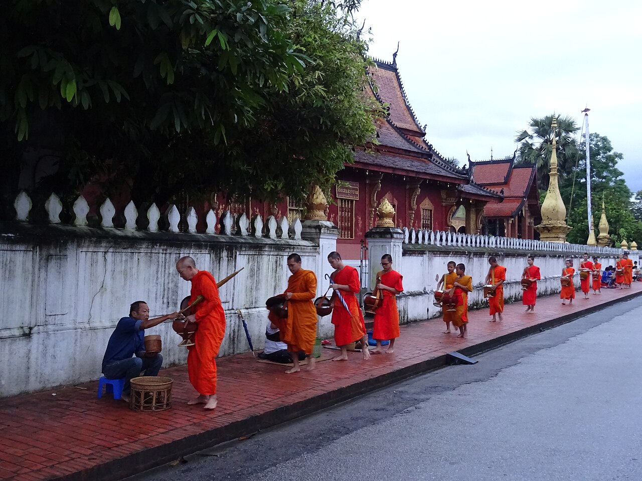 Monks in orange robes walk barefoot along a temple wall in Luang Prabang while seated almsgivers offer food at dawn.