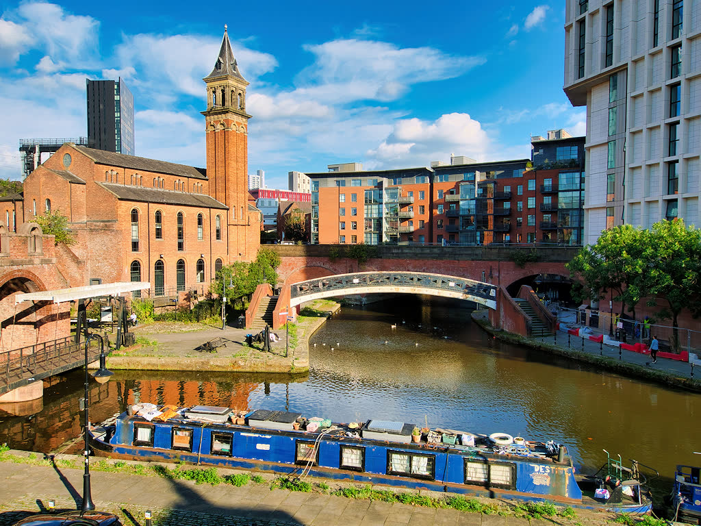 A blue narrowboat floats in Castlefield basin below a white footbridge, brick canal walls, St George's church tower, and modern Manchester apartment blocks.
