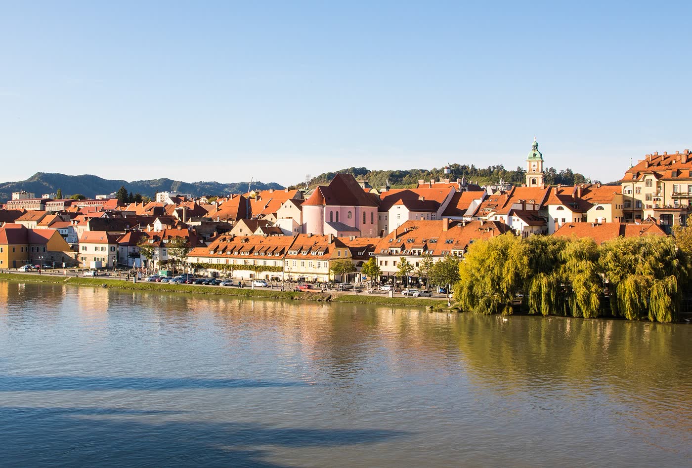 The Drava River and Lent waterfront in Maribor, with red roofs, a promenade, and the old city rising behind the river.