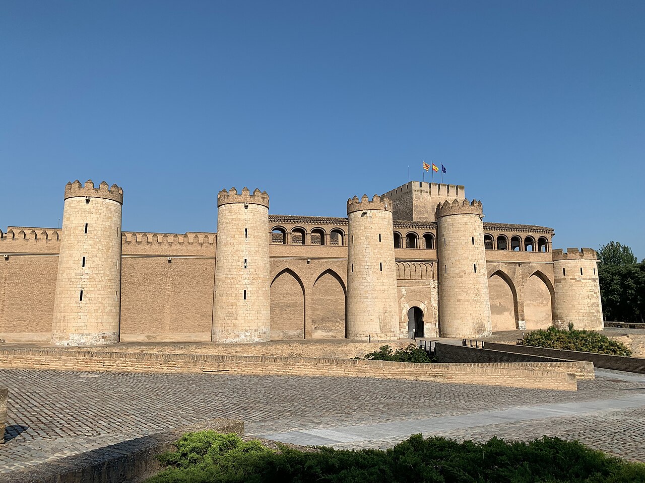 The fortified exterior of Zaragoza's Aljaferia Palace, with towers, moat-like foreground, and flags visible above the walls.