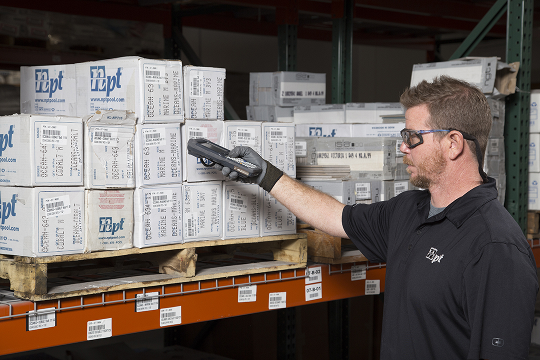 A POOLCORP worker scans boxed pool tile inventory on warehouse shelving.
