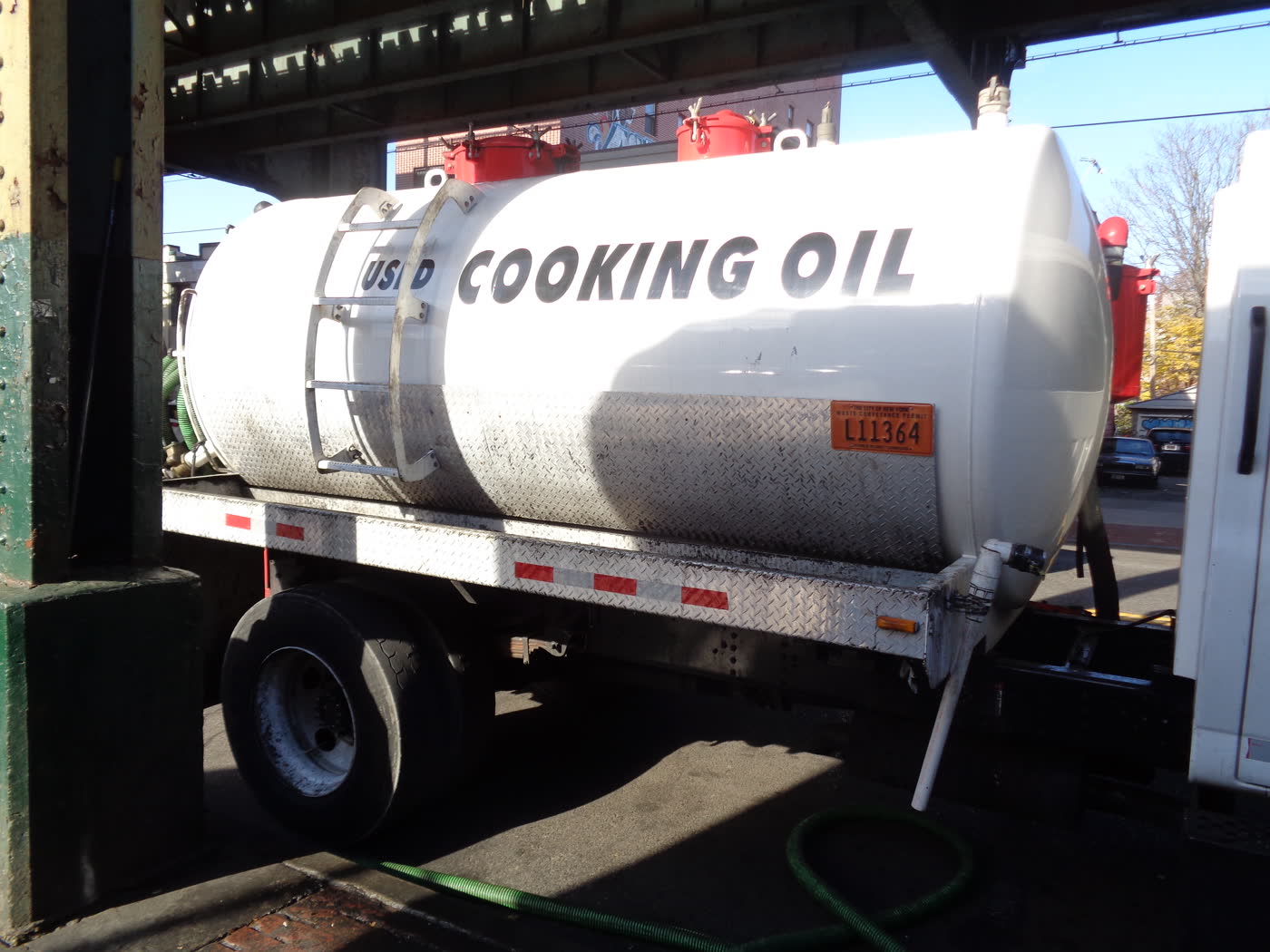 A white tanker truck marked used cooking oil parked under an elevated railway in Queens.