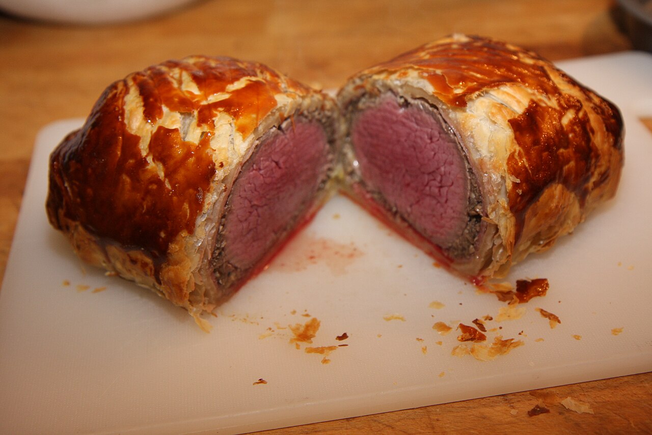 A sliced Beef Wellington showing rare beef, mushroom layer, and browned puff pastry on a cutting board.