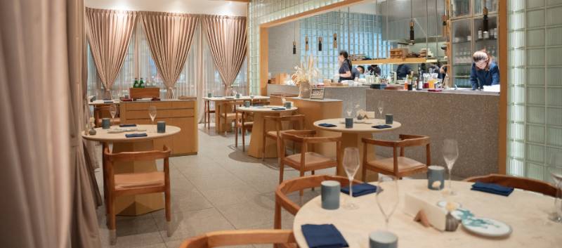 Gēn's pale-toned George Town dining room with round tables, curtains, an open kitchen, and cooks working behind a counter.
