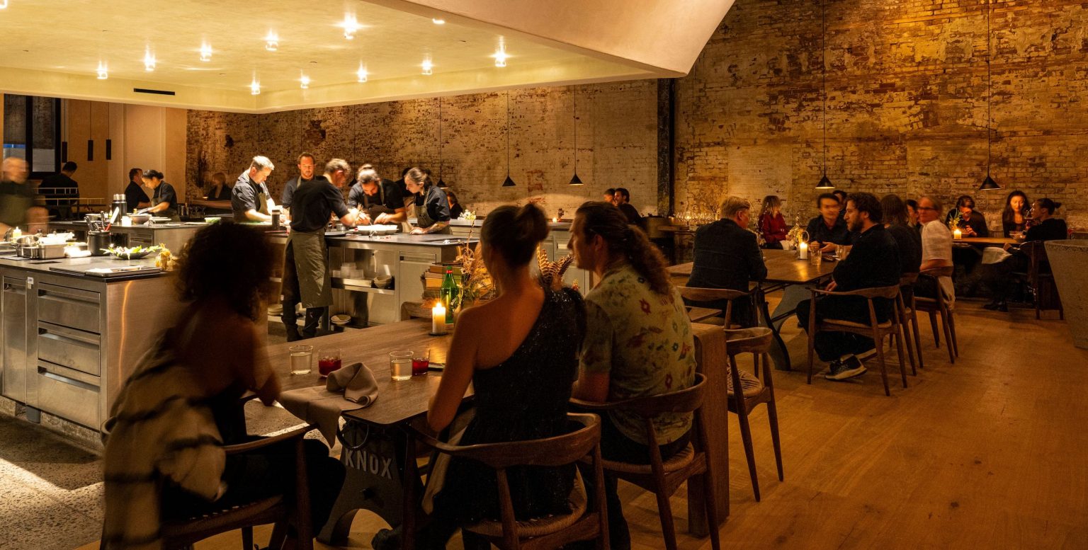 The ILIS dining room in Greenpoint, with guests seated beside a bright open kitchen under warm lights and exposed brick walls.
