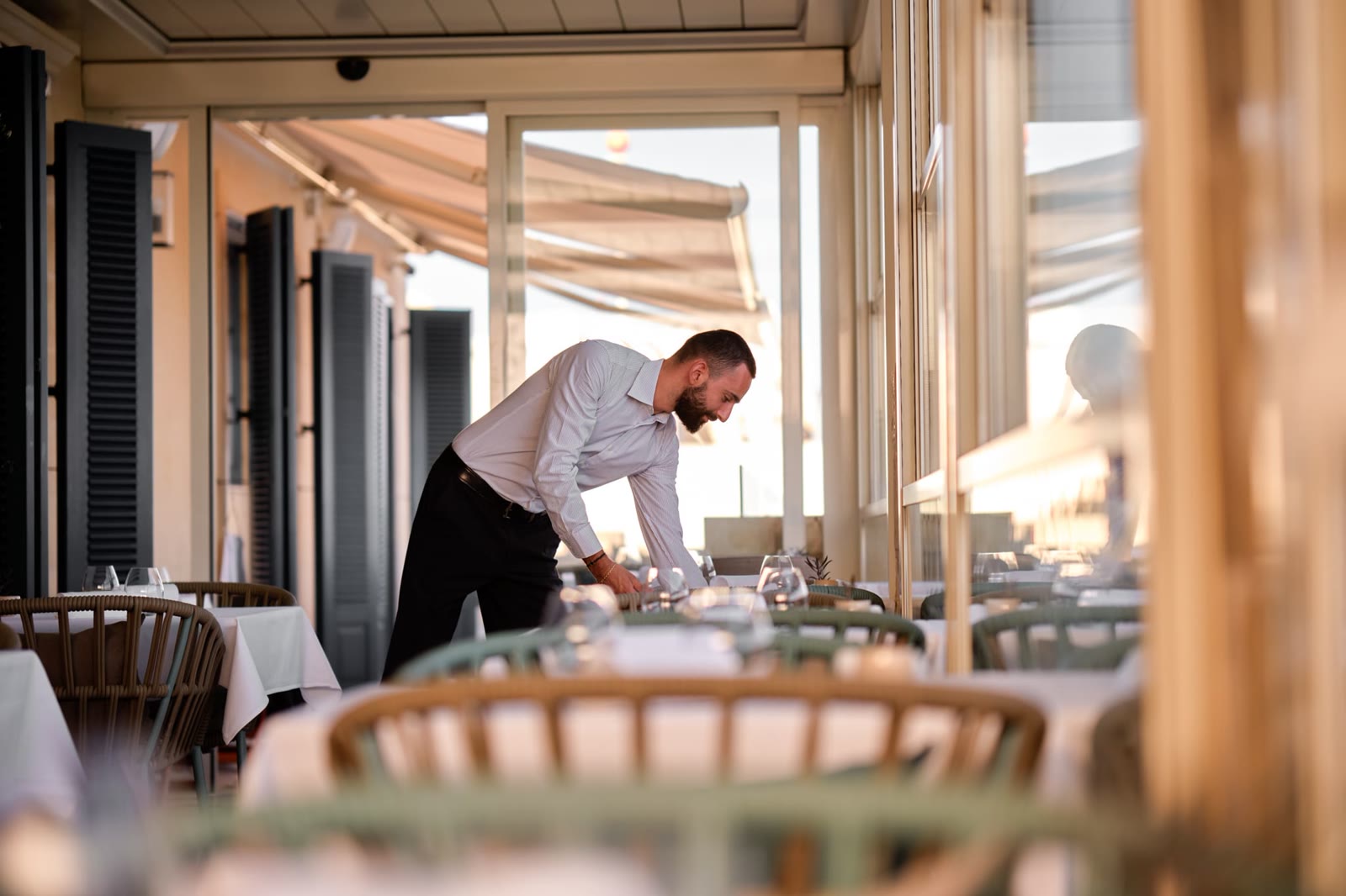 Server setting a table inside ION Harbour by Simon Rogan, with warm terrace light and glass doors along the Grand Harbour side.