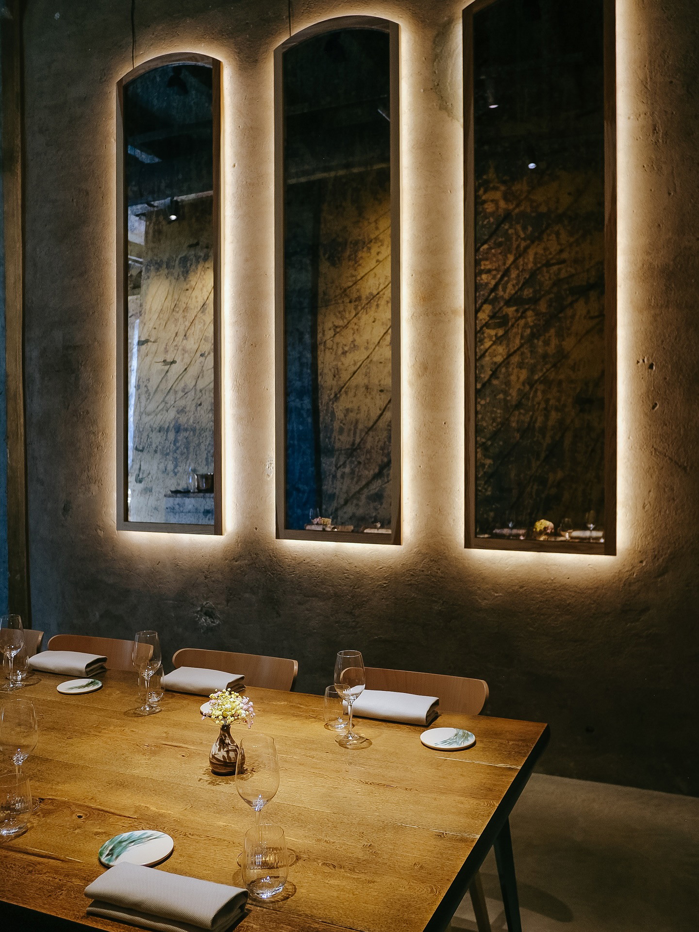Warmly lit Kíce dining room in Riga with a wooden table, set glasses, folded napkins, and tall illuminated wall mirrors.